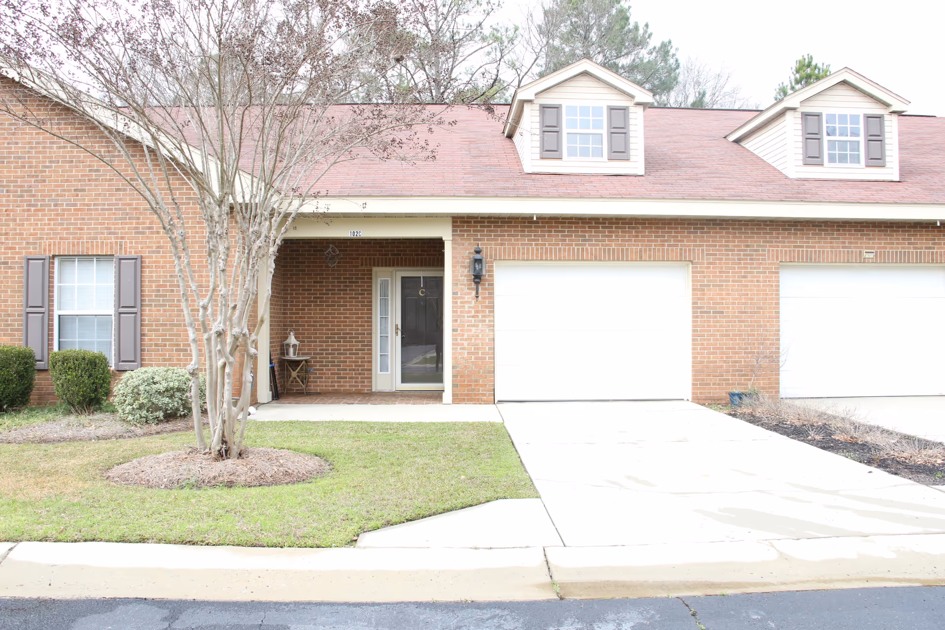 Front exterior of a brick single-story residential unit with two garage doors, a small covered porch, and a tree in the front lawn.