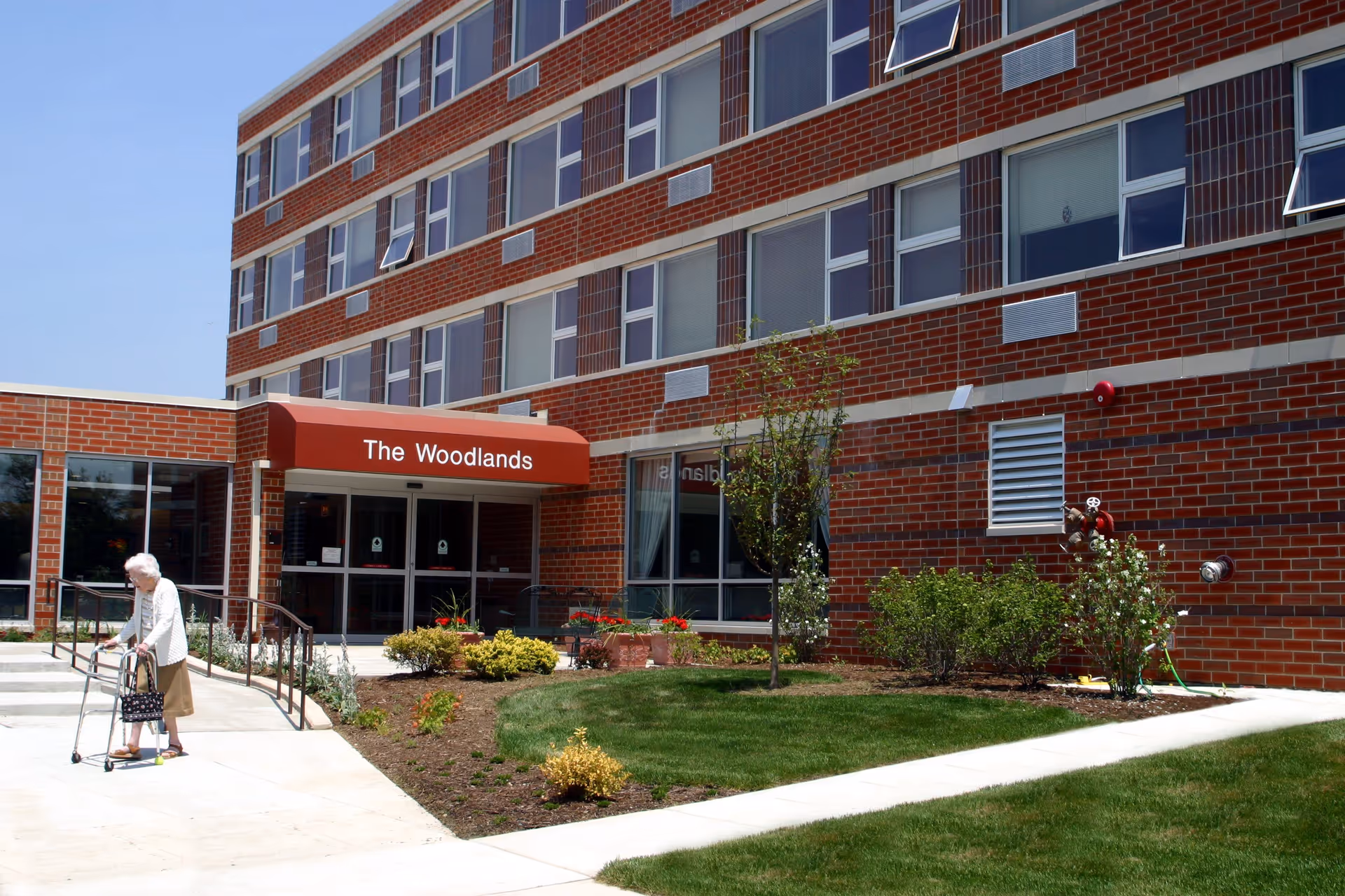 Exterior view of a multi-story brick building with a red awning labeled 'The Woodlands' above the entrance. There is a landscaped area with grass, bushes, and small trees in front of the building. An elderly woman using a walker is walking on the sidewalk near the entrance.