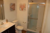 A bathroom with a white sink and countertop on a dark wood vanity, a toilet, a glass-enclosed shower with sliding doors, two framed coral-themed artworks on the wall, and a blue bath mat on the floor.
