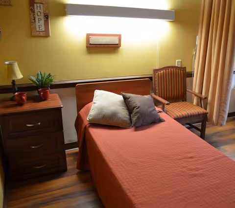 A single bed with a red bedspread and two pillows, one beige and one gray, in a room with wooden flooring. Next to the bed is a wooden nightstand with a small lamp, a plant, and two red cups. A striped armchair is positioned near the bed, and the walls are painted yellow with a decorative sign that says 'HOPE'. A curtain is partially visible on the right side of the image.