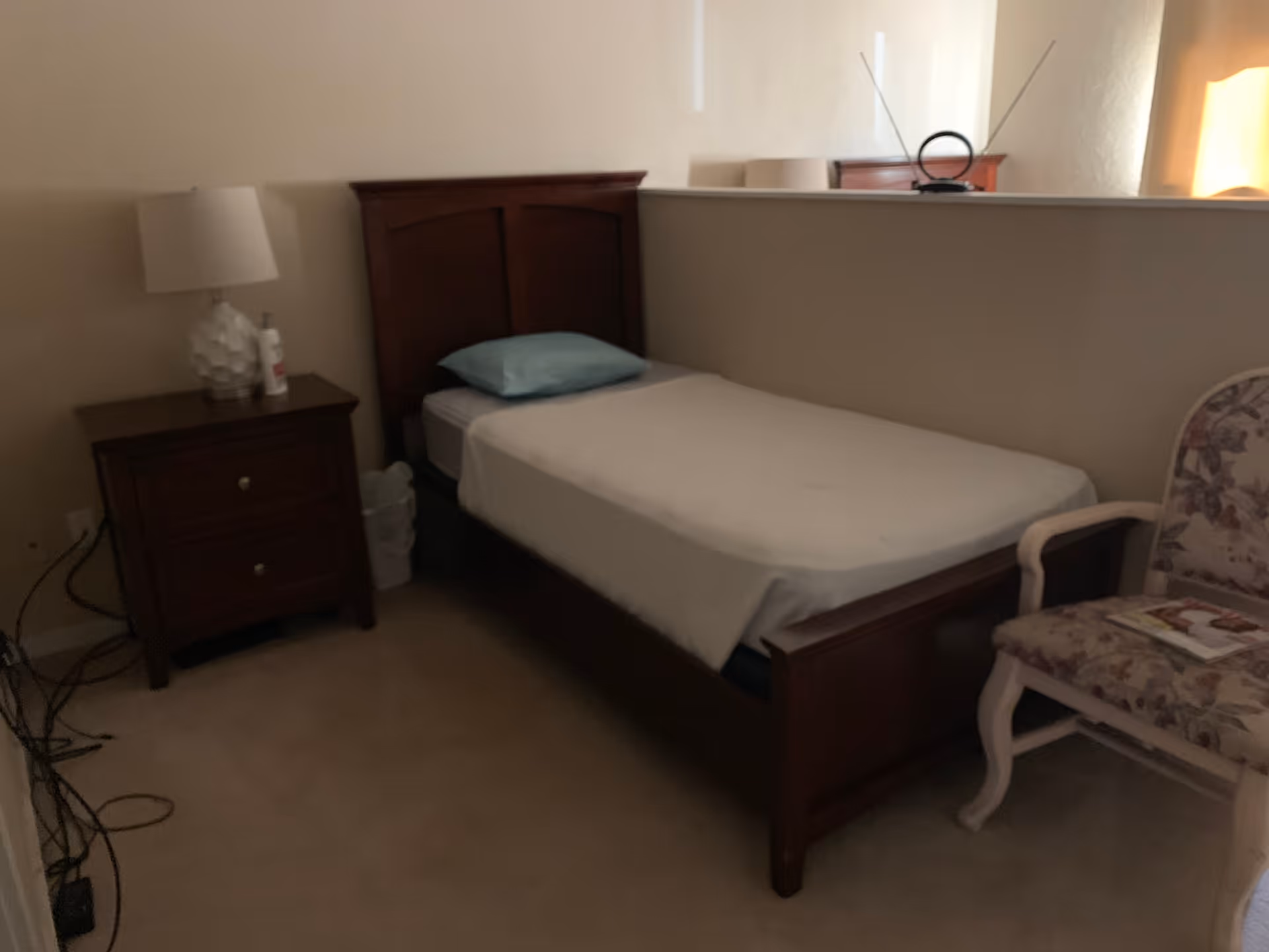 A small bedroom with a single wooden bed with a light blue pillow and white sheet. Next to the bed is a wooden nightstand with a white lamp and a bottle on it. There is a floral upholstered chair with a magazine on the seat. The room has beige carpet and a half wall separating the bed area from another space.