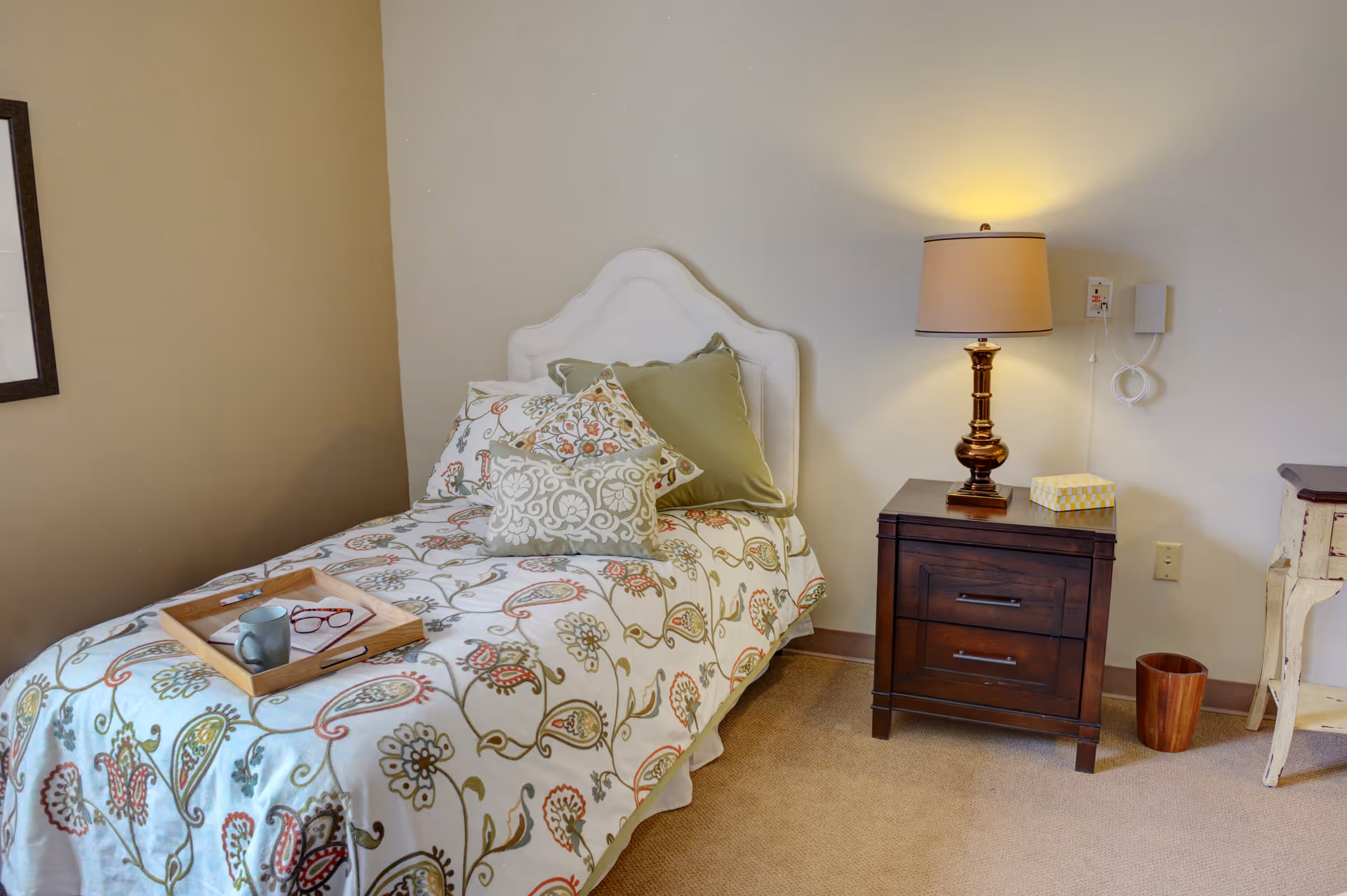 A neatly made single bed with patterned bedding and pillows beside a wooden nightstand with a lamp in a small bedroom.