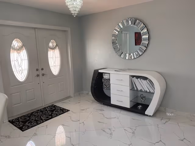 Entryway with double doors featuring oval glass inserts, a black and white modern console table with drawers and decorative items, a round silver-framed mirror on the wall, and a chandelier hanging from the ceiling. The floor is covered with white marble tiles with gray veining.