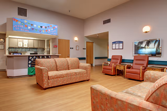 A common area in a senior living facility featuring patterned upholstered sofas and two red recliner chairs with wooden armrests. There is a reception desk with a colorful banner above it and a hallway with doors leading to other rooms. The walls are light-colored with wall-mounted lights and a framed picture of a tropical beach scene.
