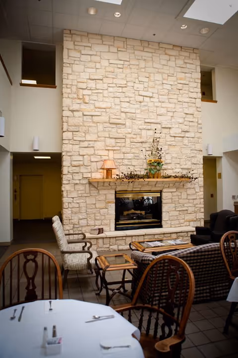 Interior view of a senior living facility lounge area featuring a tall stone fireplace with a wooden mantel decorated with a lamp and a flower arrangement. The room includes various seating options such as cushioned armchairs and wooden chairs around tables, with a tiled floor and a high ceiling with recessed lighting.
