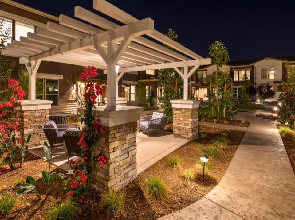 Nighttime outdoor view of a garden area at a senior living facility featuring a white pergola with stone pillars, surrounded by flowering plants and greenery. Comfortable outdoor seating is arranged under the pergola, and a well-lit pathway curves through the landscaped grounds with residential buildings visible in the background.