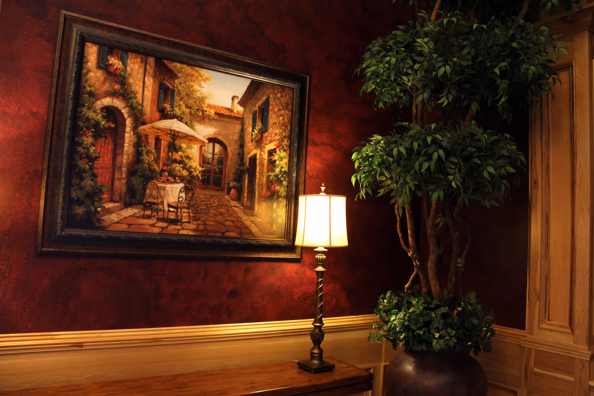 A framed painting on a red textured wall next to a lit table lamp and a large potted plant with wood paneling.