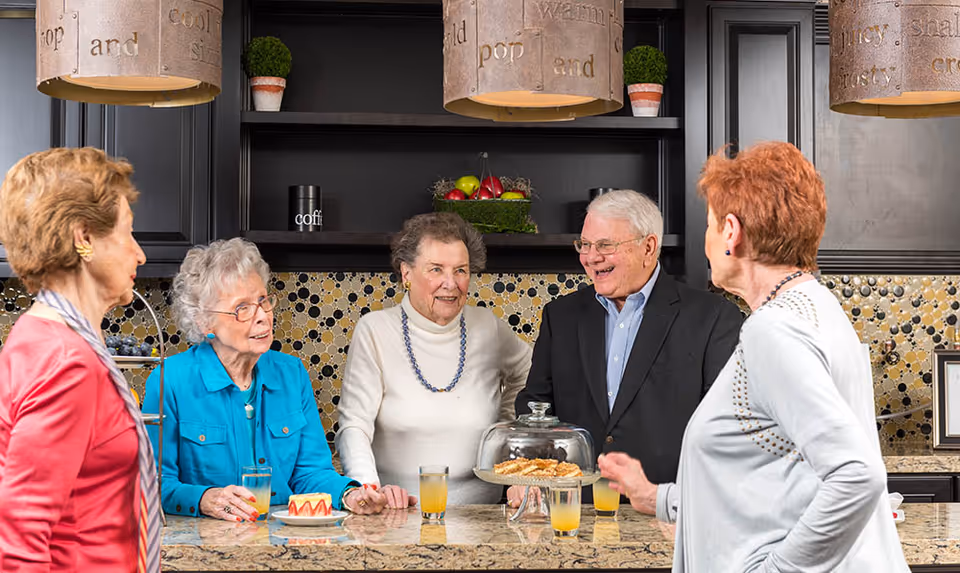 Five elderly people, four women and one man, standing around a kitchen island countertop with drinks and pastries, engaging in conversation in a well-lit kitchen with dark cabinets and decorative pendant lights.