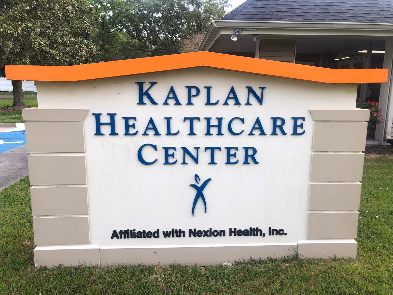 Outdoor sign for Kaplan Healthcare Center with an orange top border and beige pillars on each side, located on a grassy area with trees and a building in the background.
