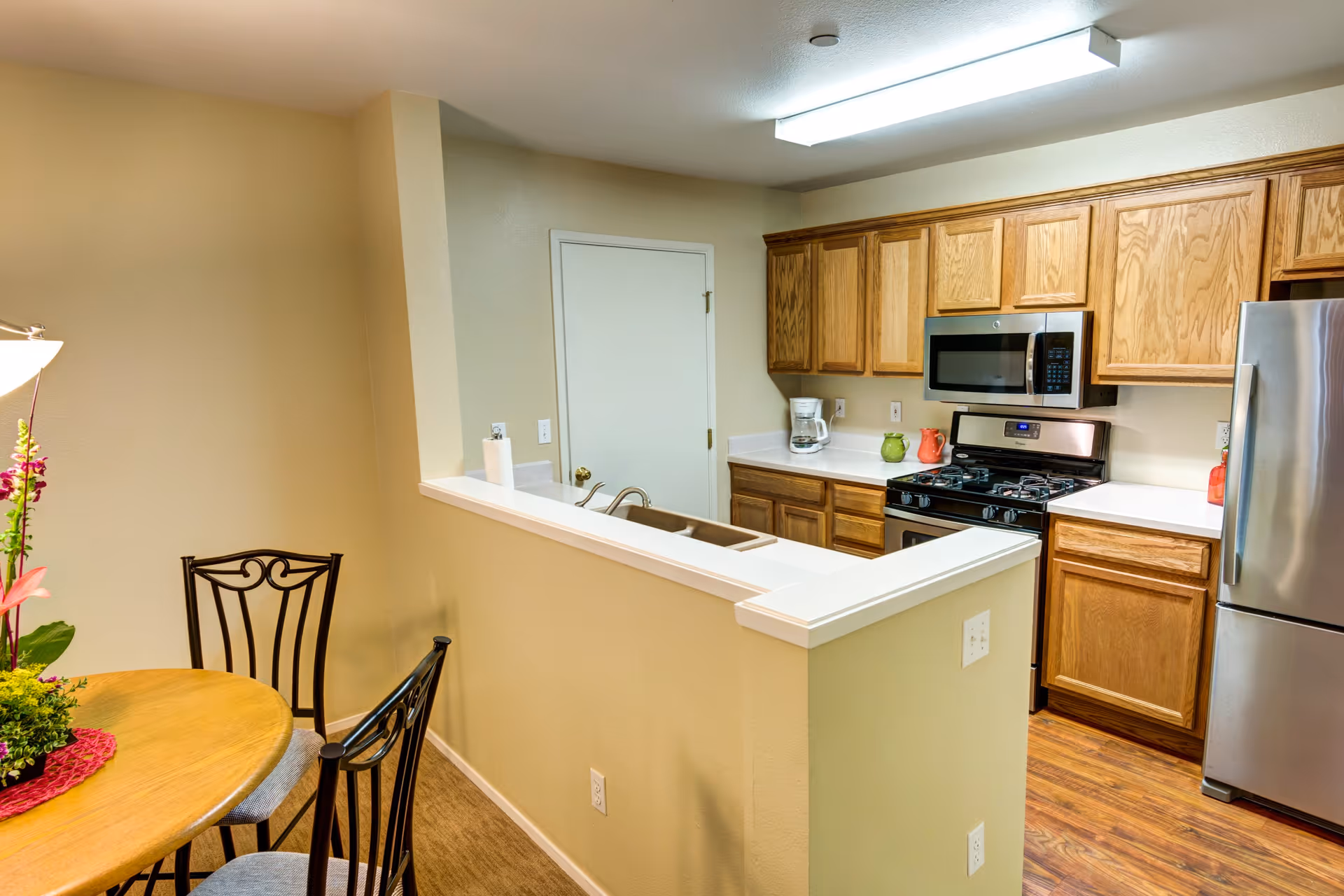 Interior view of a kitchen and dining area in a retirement community. The kitchen features wooden cabinets, a stainless steel refrigerator, a microwave, a gas stove, and a coffee maker on the countertop. The dining area has a round wooden table with black metal chairs and a floral centerpiece. The walls are painted beige, and the floor has a wood finish.