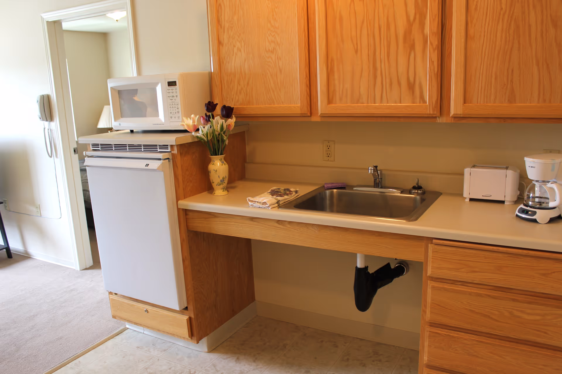 Compact kitchen with a stainless steel sink, wooden cabinets, a microwave atop a small refrigerator, and countertop appliances.