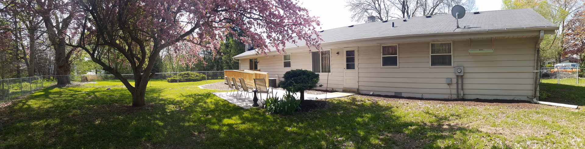 A single-story beige building with several windows and a door, surrounded by a grassy yard with a blooming tree and a small patio area with chairs. The scene is outdoors on a sunny day with clear skies.