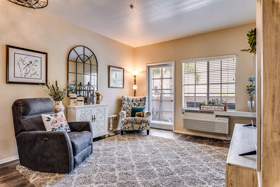 Sunlit living room with a reclining chair, patterned armchair, decorative cabinet, and large windows.