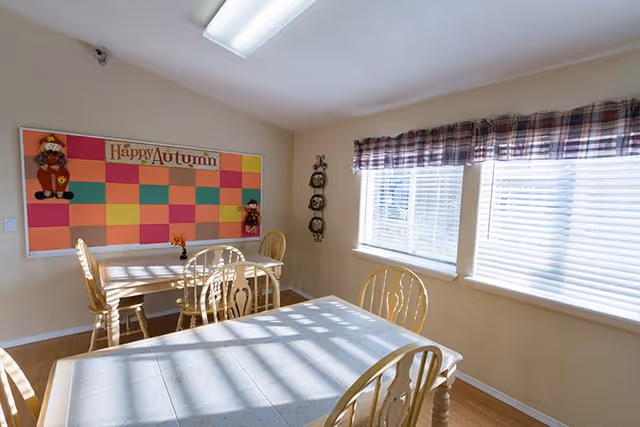 Sunlit dining/activity room with wooden tables and chairs, a 'Happy Autumn' bulletin board, and windows with blinds.