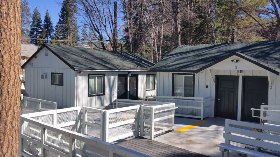 Front exterior of single-story white cottages with dark green roofs, numbered entrance doors and accessible ramps amid tall trees.