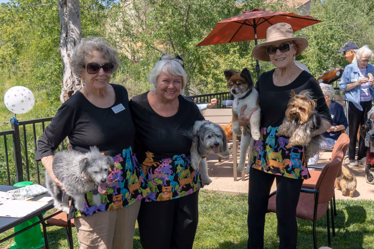 Three elderly women standing outdoors on grass, each holding a small dog. They are smiling and wearing matching black tops with colorful dog-themed aprons. Behind them, there are other people and outdoor furniture, with trees and greenery in the background.