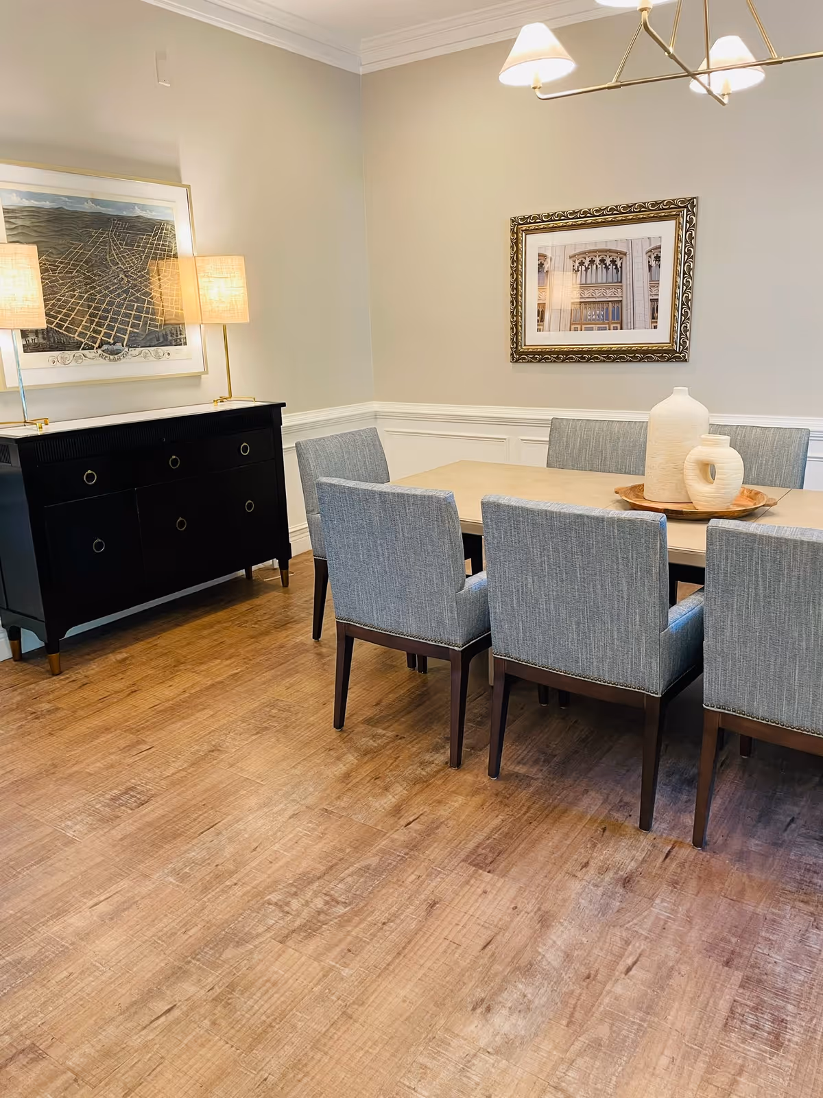A dining room with a rectangular wooden table surrounded by six upholstered chairs in a light gray fabric. On the table, there is a wooden tray holding two decorative vases. The room has light-colored walls with white wainscoting and crown molding. A black sideboard with two lamps and a framed map hangs above it on one wall. Another framed picture is hung on the adjacent wall. The floor is wood with a natural finish, and a modern chandelier with three lamps hangs above the table.