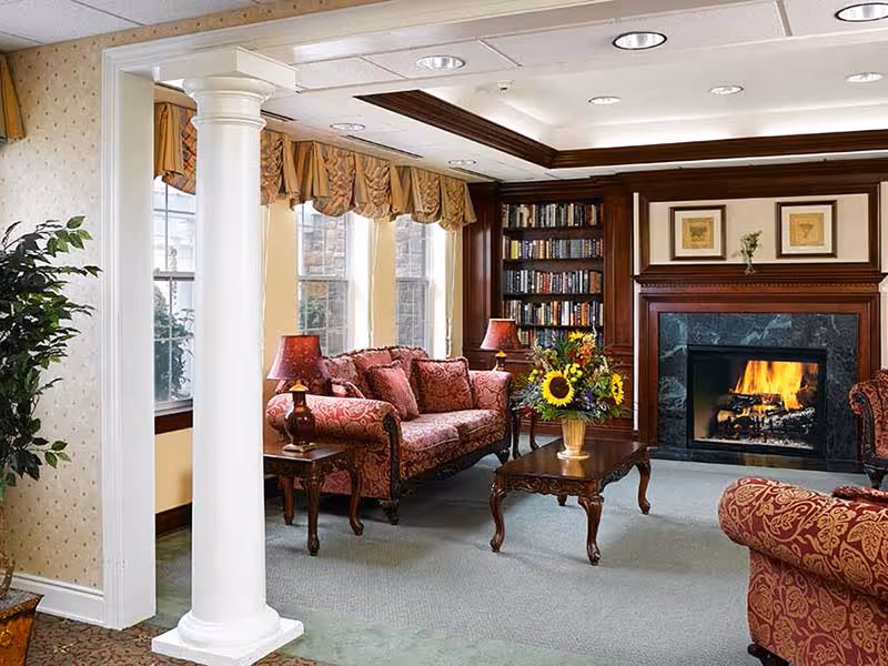 A cozy living room with classic decor featuring a red patterned sofa and armchair, a wooden coffee table with a vase of sunflowers, a lit fireplace with a dark marble surround, built-in wooden bookshelves filled with books, and large windows with beige curtains allowing natural light to enter.