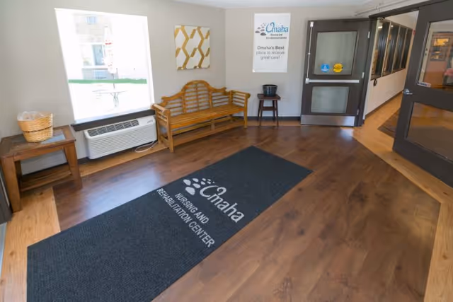 Entrance lobby with a wooden bench, decorative wall art, and a welcome mat reading 'Omaha Nursing and Rehabilitation Center' in front of glass entry doors.