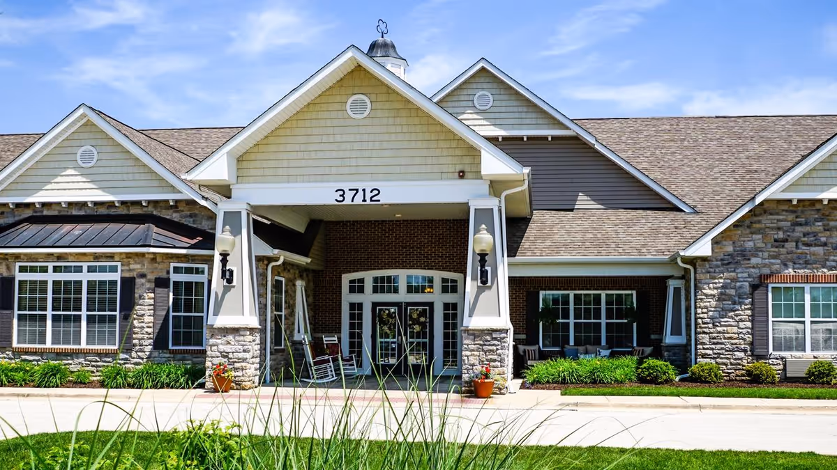 Front exterior view of a senior living facility building with stone and siding facade, large windows, a covered entrance with the number 3712 above it, and a clear blue sky in the background.