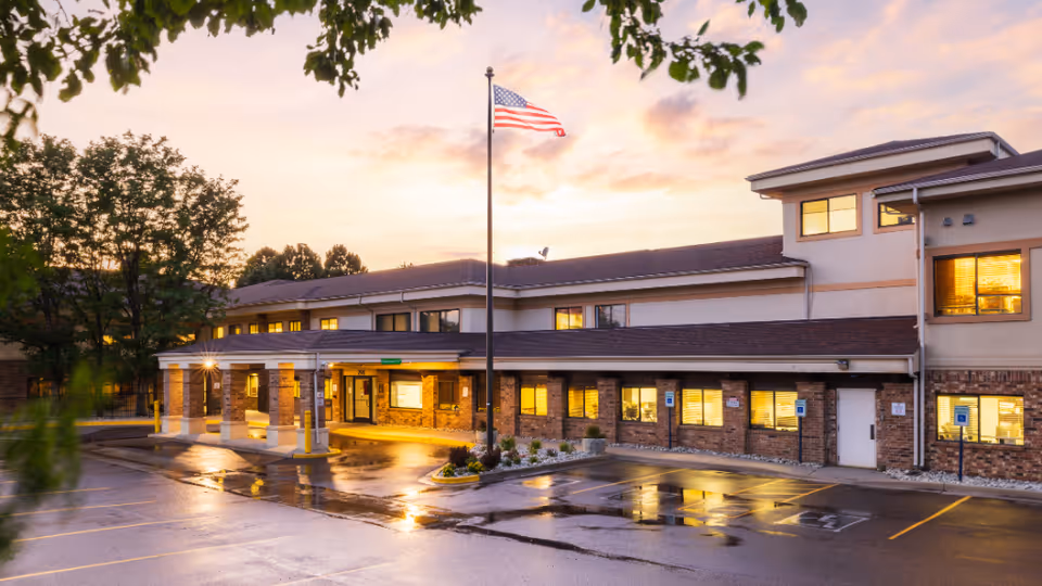 Front entrance of a senior living facility at dusk with an American flag and wet parking lot reflecting lights.