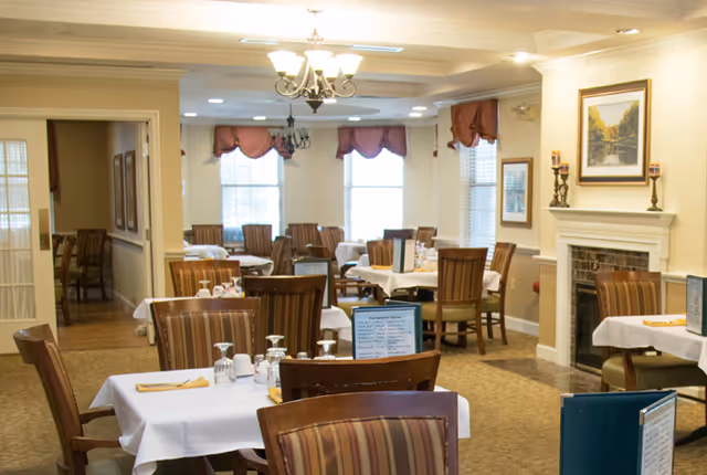 A well-lit dining room with multiple tables covered in white tablecloths, each set with glasses, napkins, and menus. The room features striped upholstered chairs, large windows with red valances, a chandelier, and a fireplace with decorative candles and framed artwork above it.
