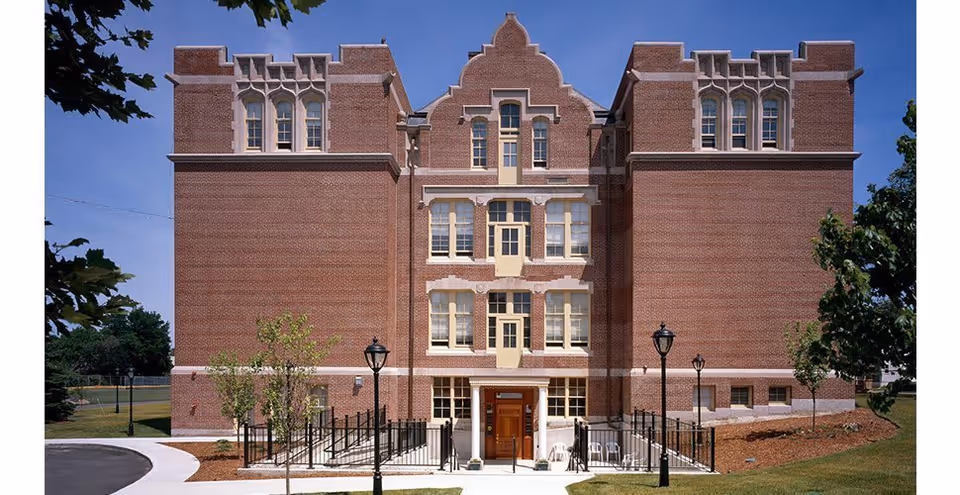 Front exterior view of a large, historic brick building with multiple windows and decorative architectural details. The entrance is centered with a small set of stairs and a ramp leading up to it, flanked by black metal railings and street lamps. There are small trees and landscaped areas on either side of the entrance.