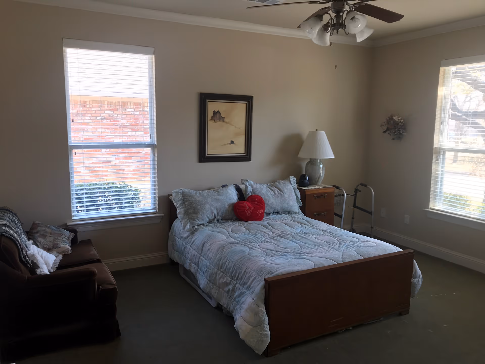 A bedroom with a bed covered in a light blue quilt and pillows, including a red heart-shaped pillow. There is a wooden nightstand with a lamp and a small black object next to it. A walker is positioned near the nightstand. The room has two windows with white blinds, a brown armchair with pillows, a ceiling fan with lights, and a framed picture on the wall.