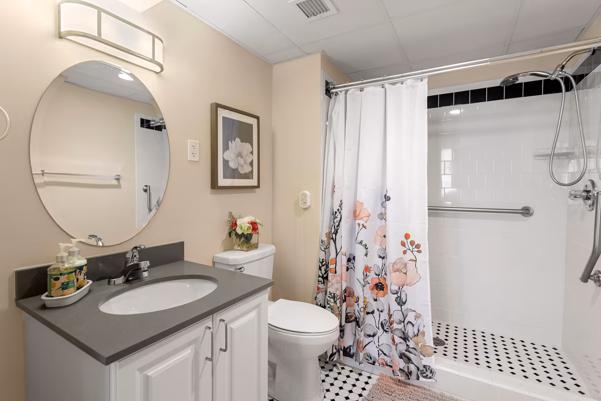 A clean bathroom with a white vanity and gray countertop, an oval mirror above the sink, a toilet with a small flower arrangement on top, and a shower with a floral-patterned curtain and grab bars inside.