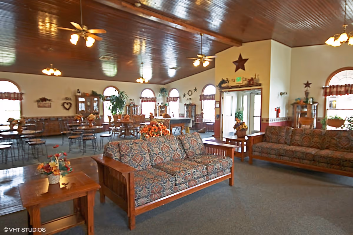 A spacious common area with wooden ceiling and ceiling fans, featuring patterned sofas, wooden side tables with flower arrangements, and multiple dining tables and chairs near arched windows with valances. The room is decorated with star-shaped wall hangings and plants.