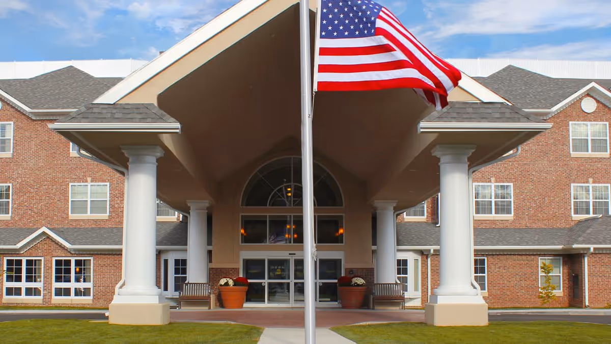 Front entrance of a brick building with large white columns supporting a covered driveway. An American flag is prominently displayed on a flagpole in the center. There are benches and large flower pots near the entrance doors.