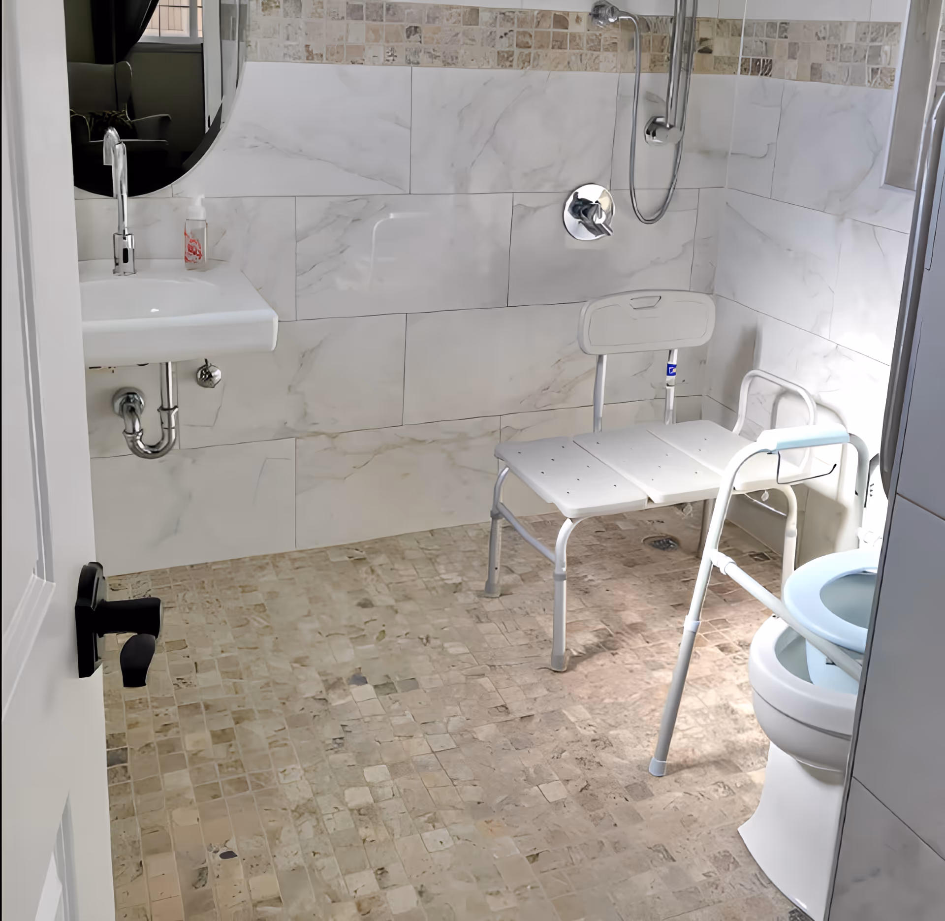 A bathroom with beige tiled floor and white marble-patterned wall tiles. It features a white sink with a chrome faucet, a round mirror above the sink, a shower area with a handheld showerhead, a white shower chair, and a toilet equipped with a white safety frame.