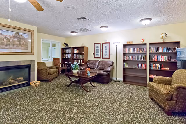 A cozy living room area with patterned carpet, a brown leather sofa, two upholstered armchairs, a wooden coffee table with a flower vase, two bookshelves filled with books, a floor lamp, a water dispenser, and a framed picture above a fireplace.