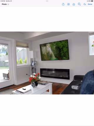 A modern living room with a wall-mounted flat-screen TV displaying a nature scene above a sleek electric fireplace. There is a white coffee table with a vase of red and white flowers and some books on it. A black leather sofa with a blue throw blanket is partially visible. Large windows and a glass door provide natural light and a view of a fenced outdoor area with greenery.
