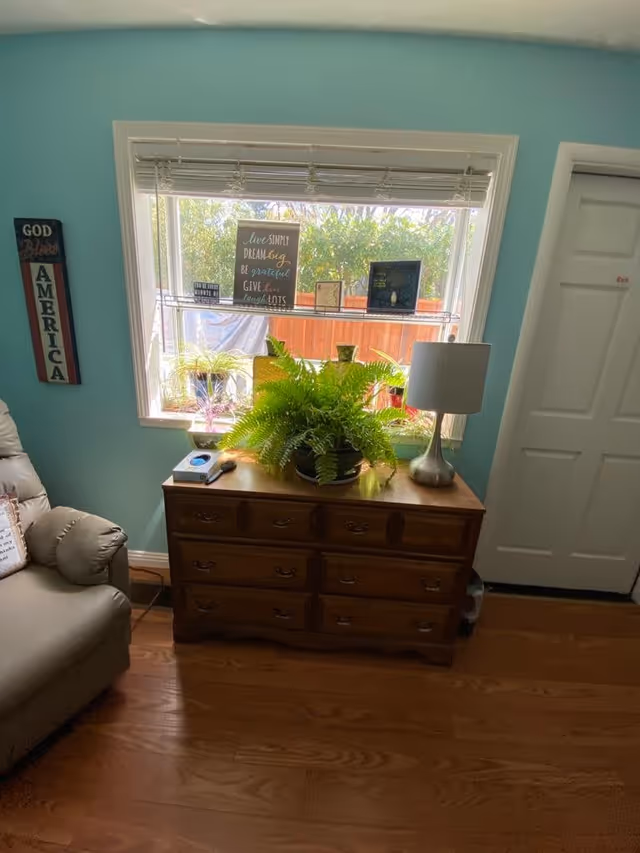 A cozy room with light blue walls featuring a window with blinds partially open, letting in natural light. Below the window is a wooden dresser with six drawers, decorated with a large green fern plant and a silver lamp. On the window sill, there are several small framed signs and plants. To the left, part of a beige recliner chair is visible, and on the wall next to the window hangs a vertical sign that reads 'God Bless America'. To the right, there is a white door with a small red label.