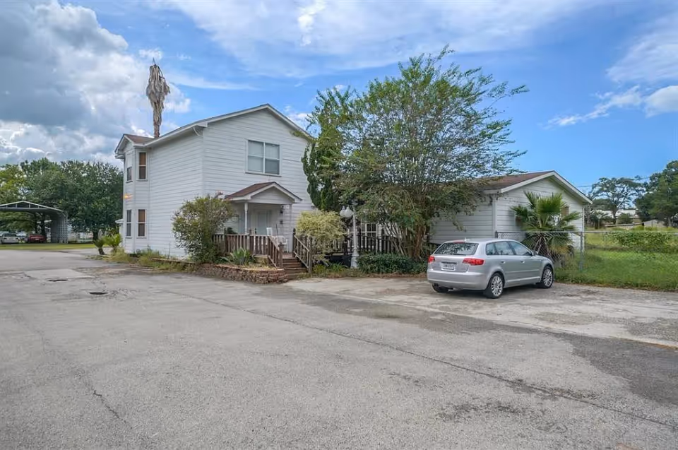 Exterior view of a two-story white building with a small porch and stairs, surrounded by trees and bushes. A silver car is parked on the paved area in front of the building. The sky is partly cloudy with patches of blue.