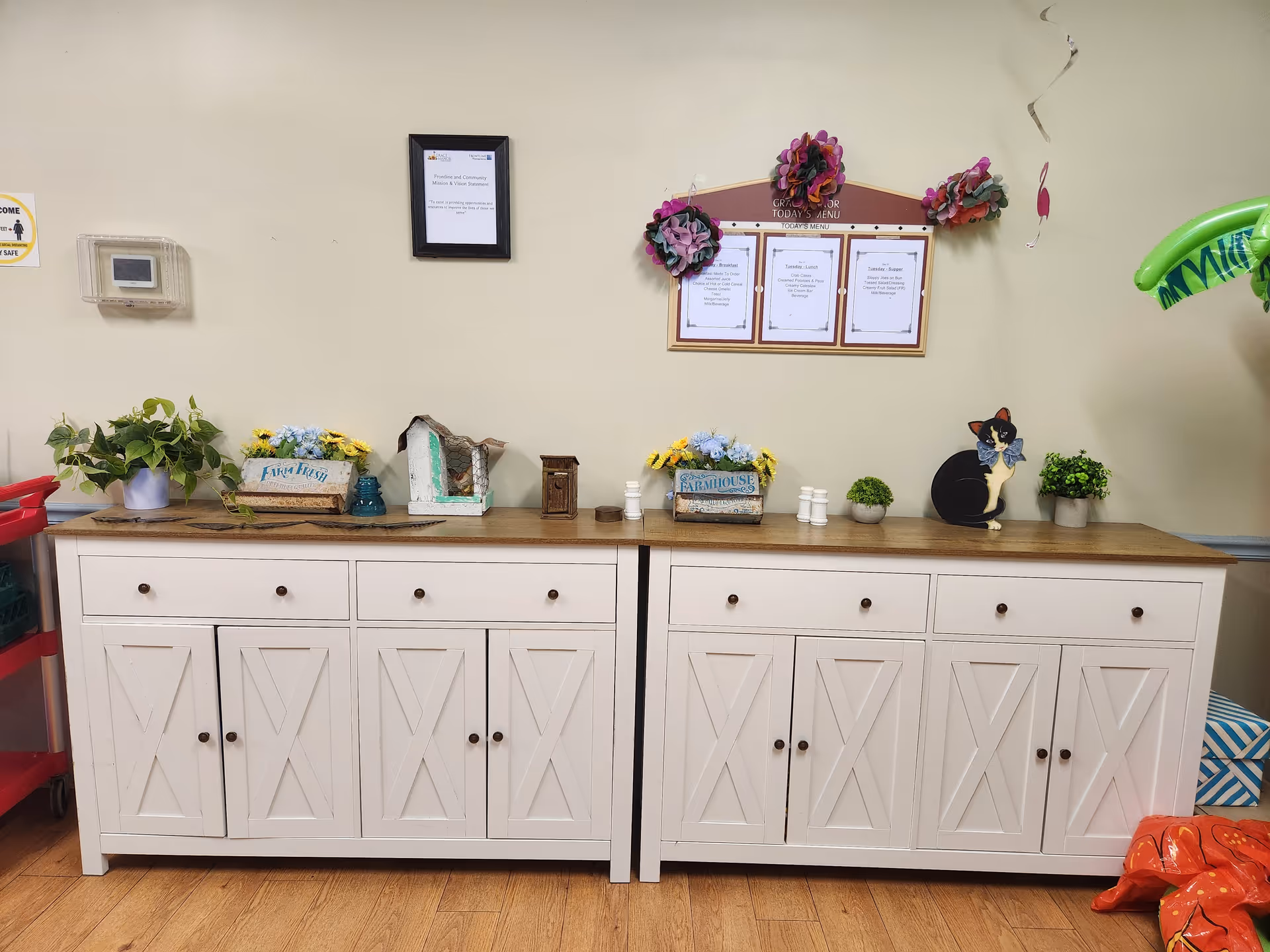 Two white wooden cabinets with brown knobs and wooden tops placed side by side against a beige wall. On top of the cabinets are decorative items including potted plants, a small birdhouse, salt and pepper shakers, and a cat figurine. Above the cabinets on the wall is a framed menu board decorated with paper flowers and a framed notice. The floor is wooden, and part of a red cart and some colorful items are visible on the sides.
