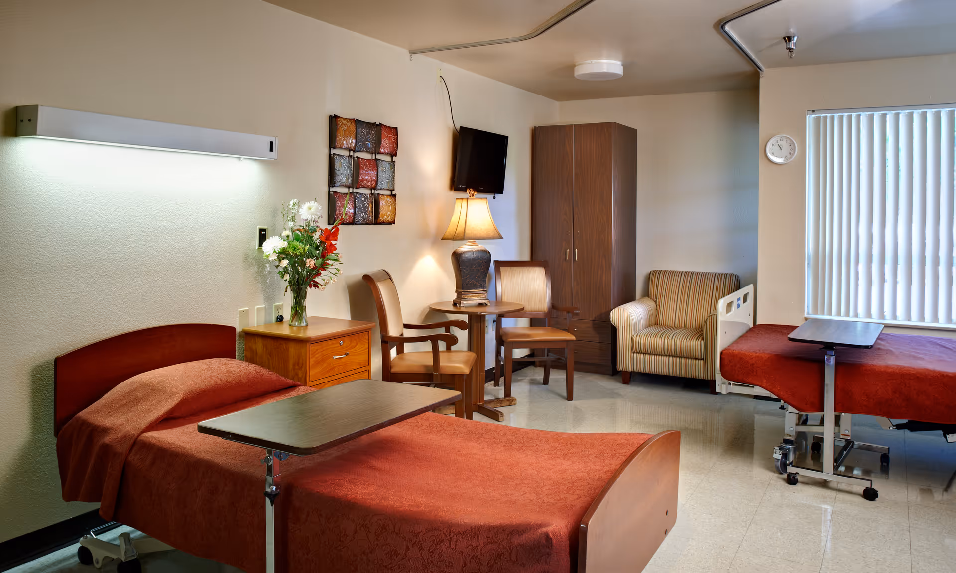 Bright senior living bedroom with two beds, a bedside table with flowers, chairs, a small table, wardrobe and wall-mounted TV.