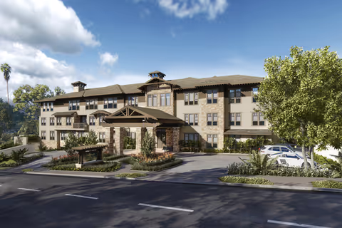 Three-story Mediterranean-style senior living building with a covered porte-cochere entrance, landscaped circular driveway, and parked cars under a blue sky.