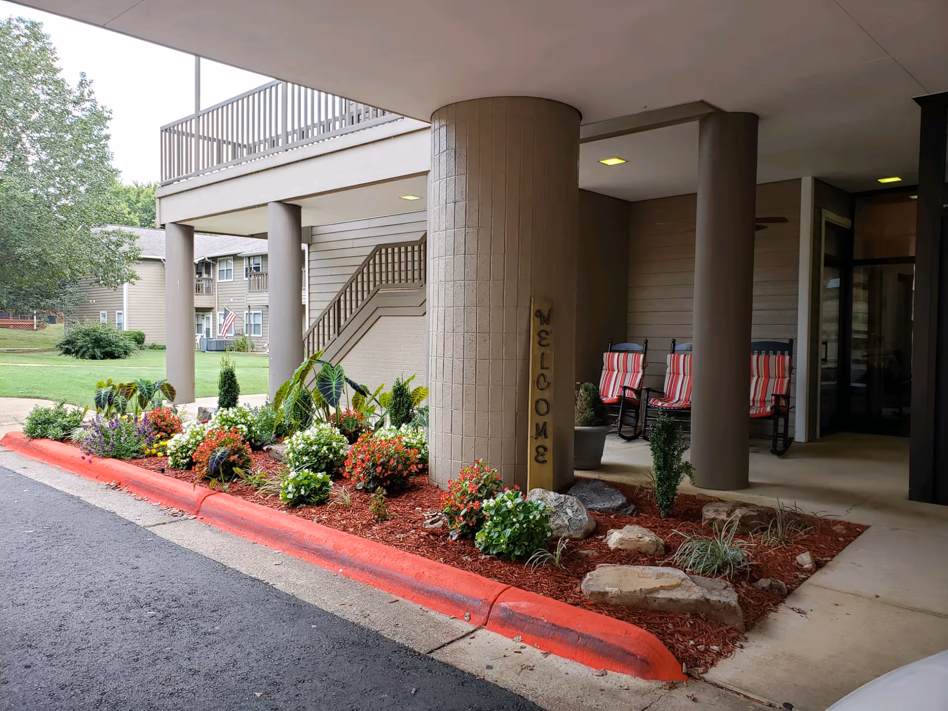 Entrance area of a retirement community building with a covered driveway supported by large columns. There is a landscaped garden bed with various flowers and plants bordered by a red-painted curb. A vertical wooden sign on one column reads 'WELCOME'. In the background, there are rocking chairs with red and white striped cushions on a porch area, and a residential building with an American flag is visible across a grassy lawn.