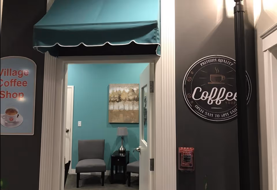 Interior view of a coffee shop area with a teal awning above the entrance. Inside, there are two gray chairs with a small black table and a lamp between them, against a teal wall with a painting of trees. On the right wall, there is a round sign that reads 'Premium Quality Coffee Shop Open 5AM to 6PM Daily'. On the left wall, a sign partially visible reads 'Village Coffee Shop'.