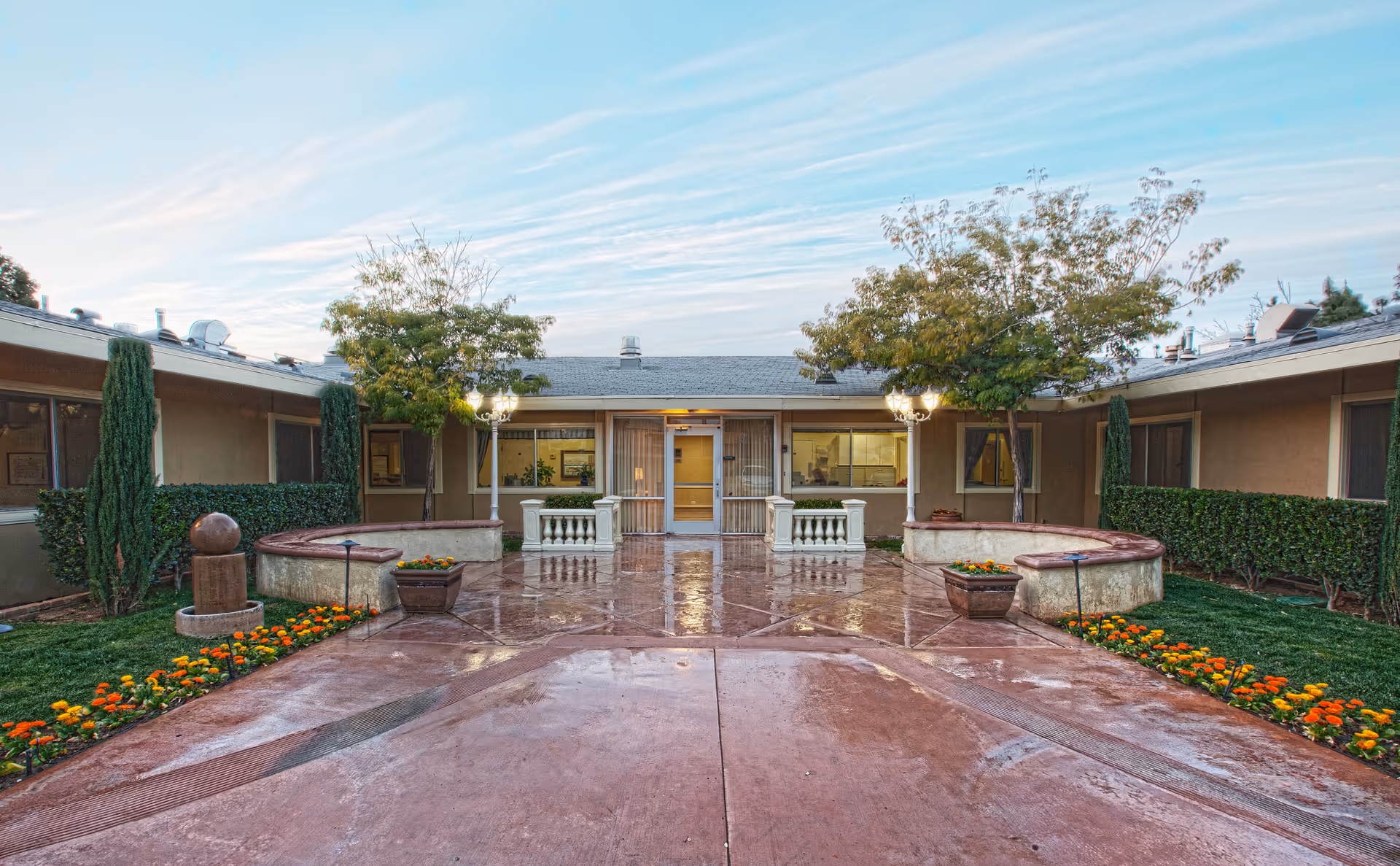 Outdoor courtyard area of a senior living facility with a wet concrete pathway, flower beds with orange and yellow flowers, two trees, and a building entrance with large windows and a door in the background.
