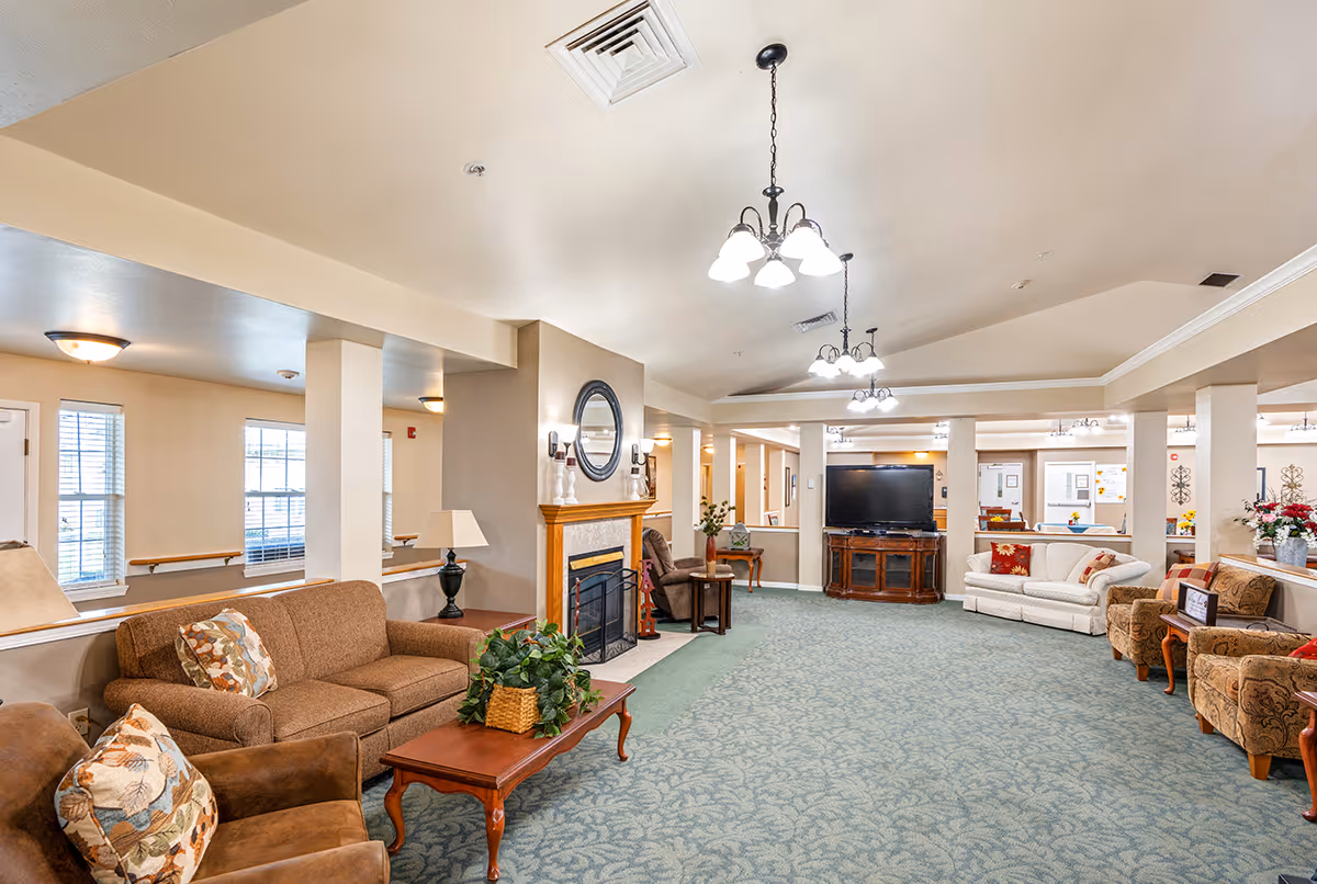 A spacious senior living facility common area with multiple seating arrangements including sofas and armchairs. The room features a fireplace with a round mirror above it, a large flat-screen TV on a wooden stand, and several light fixtures hanging from the ceiling. The carpet is patterned in blue-green tones, and the walls are painted beige. There are windows allowing natural light to enter the room.