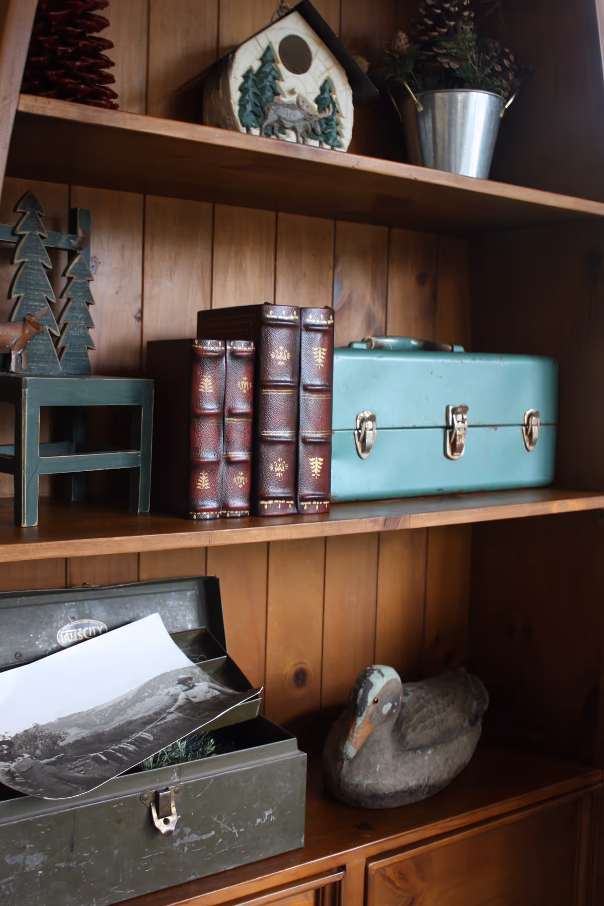 Wooden shelves with decorative items including a small wooden chair with tree cutouts, a birdhouse, a metal bucket with pinecones, three leather-bound books, a light blue metal box with latches, an open green metal box containing a black and white photograph, and a carved wooden duck.