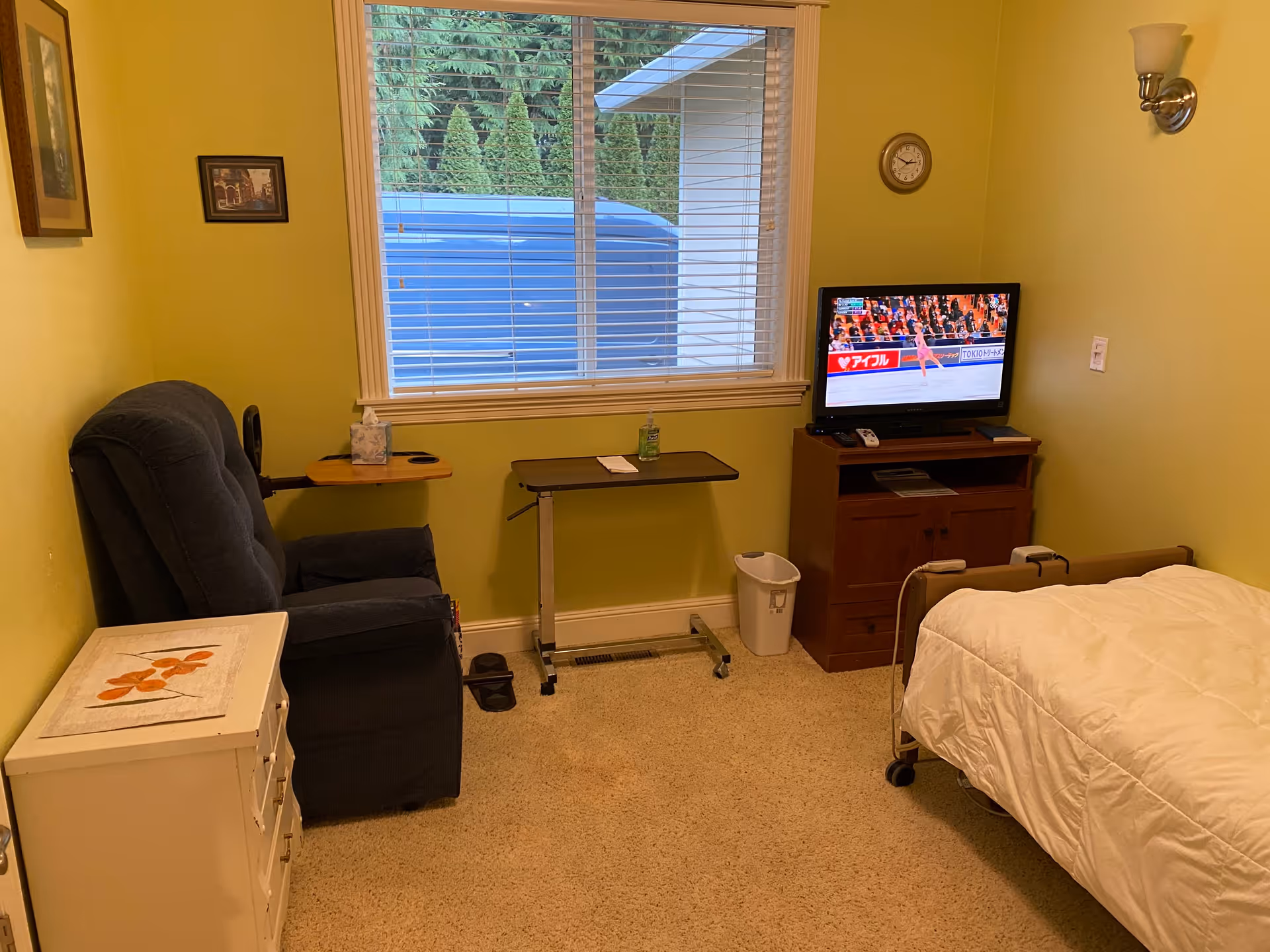 A small bedroom with light green walls featuring a single bed with white bedding, a dark blue recliner chair with an attached wooden tray, a small white dresser with a floral design on top, a rolling overbed table, a wooden TV stand with a television showing a figure skating event, a wall clock, and a window with white blinds overlooking greenery outside.
