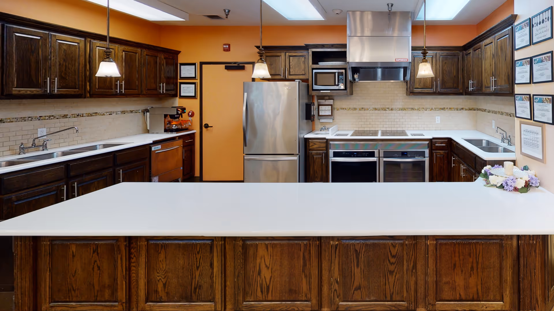 A spacious kitchen with dark wooden cabinets, white countertops, and stainless steel appliances including a refrigerator, microwave, and double ovens. The walls are painted orange, and there are pendant lights hanging over the counter. A small floral arrangement with a candle is placed on the right side of the counter. Several framed certificates or documents are displayed on the wall to the right.