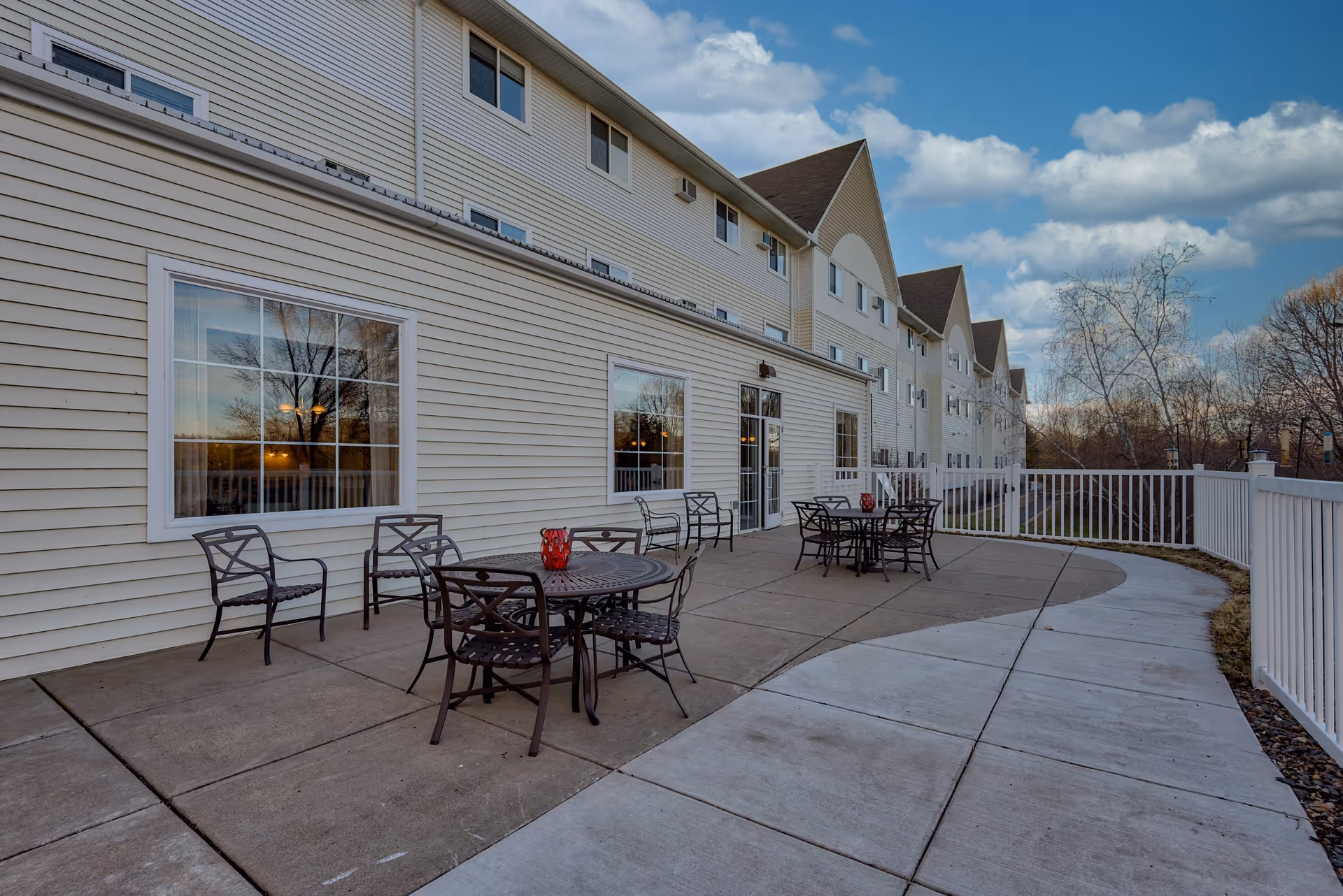 Outdoor patio area at Amira Choice Champlin with several metal tables and chairs arranged on a concrete surface next to a light-colored building. The patio is enclosed by a white fence and there are trees and a partly cloudy sky in the background.