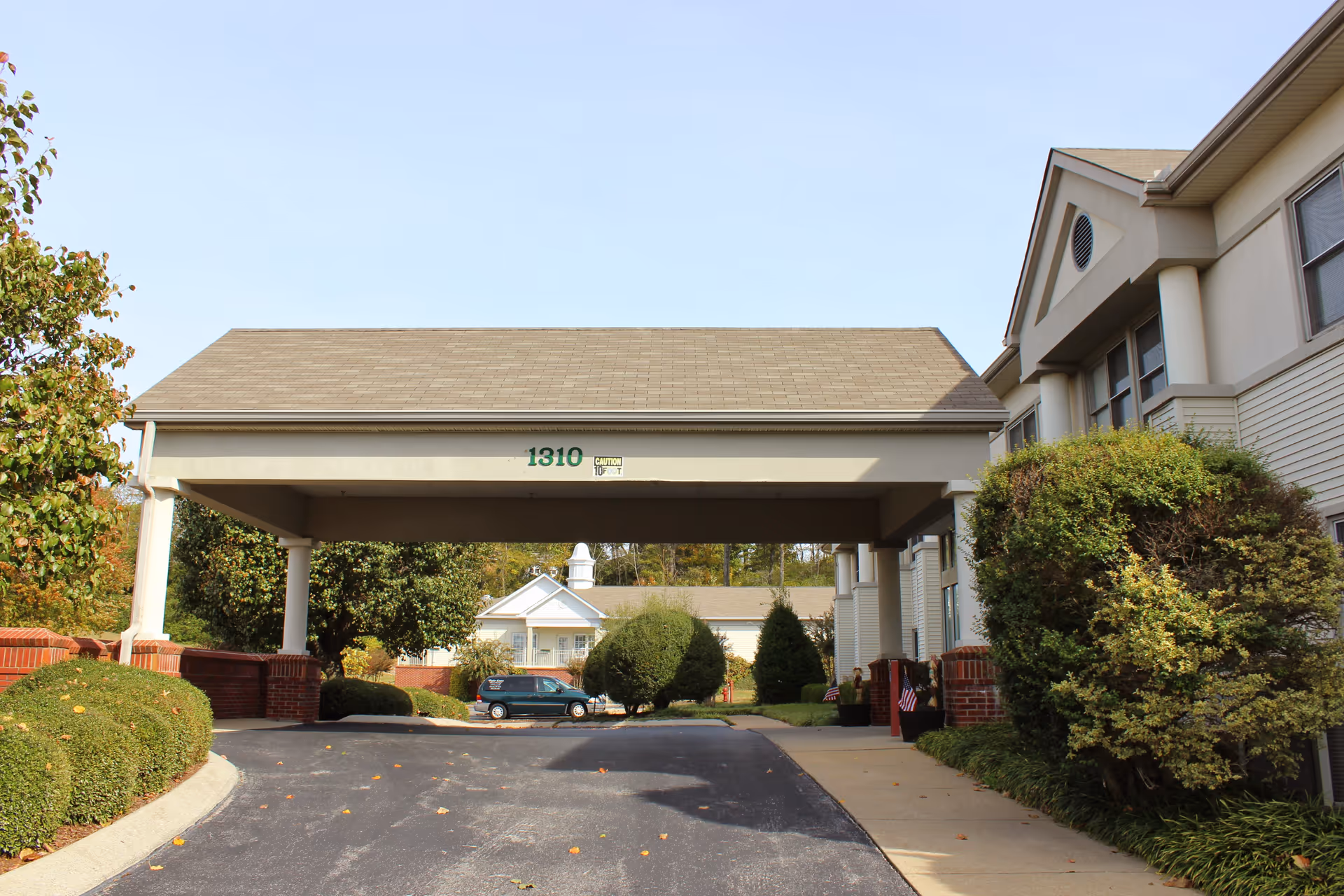 Entrance driveway of Poplar Estates Senior Living Community with a covered drop-off area supported by white columns, surrounded by trimmed bushes and trees, and a building with beige siding and multiple windows.