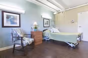 A nursing-home patient room with a single hospital bed, nightstand and lamp, upholstered chair, framed artwork, and wood-look flooring.