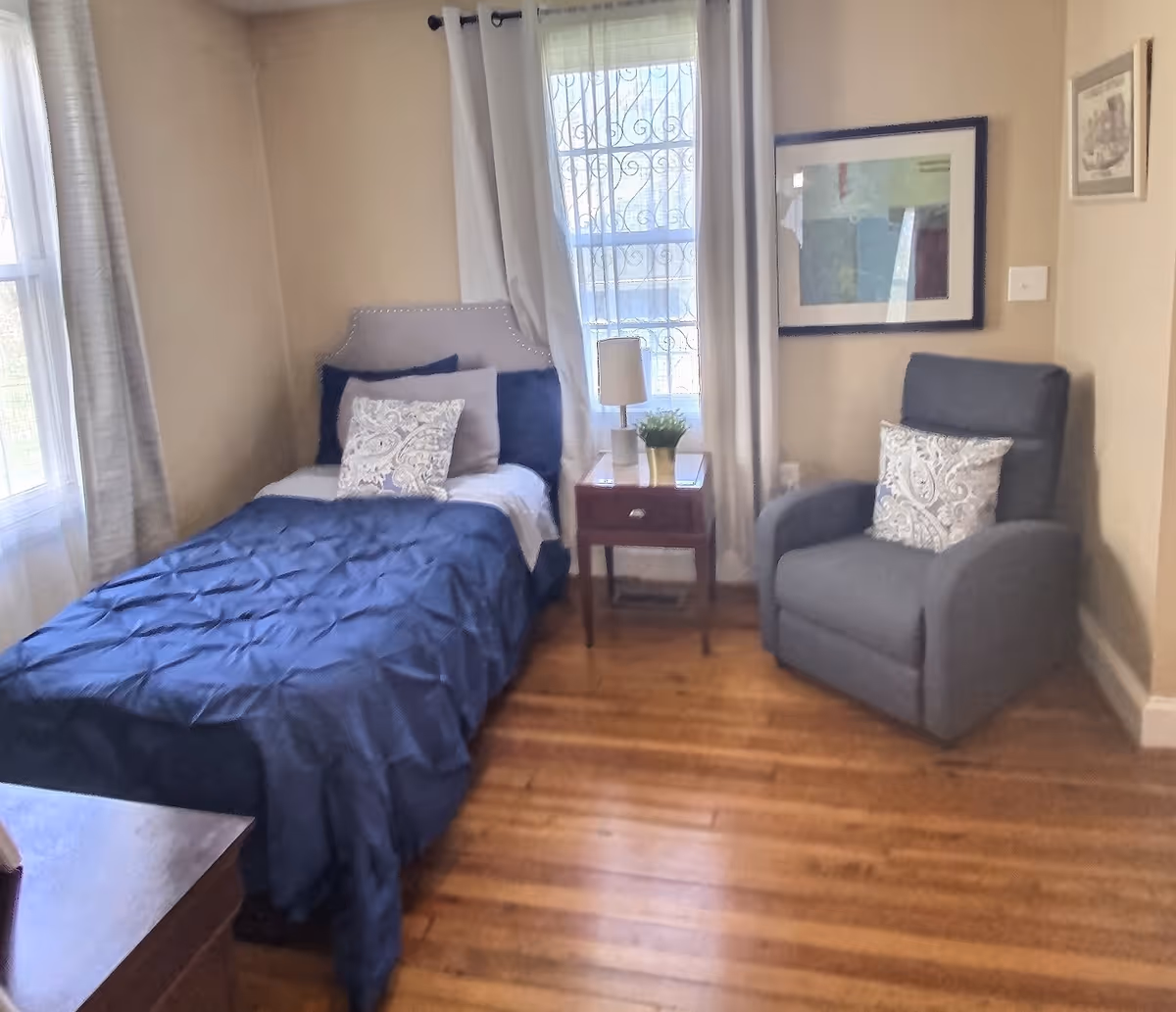 A cozy bedroom with a single bed covered in a dark blue comforter and decorative pillows. Next to the bed is a wooden nightstand with a small lamp and a potted plant. A comfortable gray armchair with a patterned pillow is placed against the wall. The room has wooden floors, beige walls, two windows with white curtains, and framed artwork on the walls.